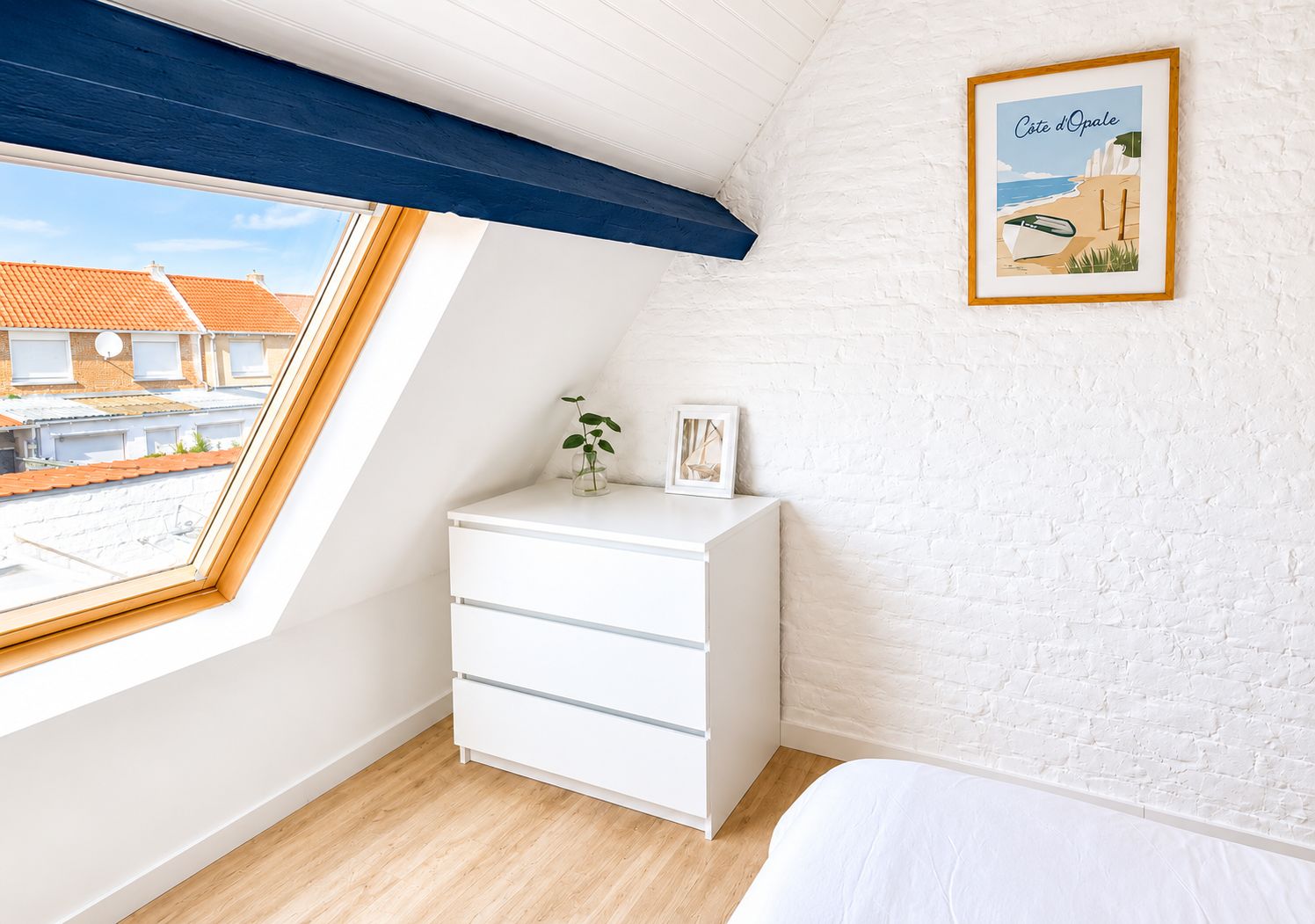 Een slaapkamer met dakraam, een witte dressoir, een bed en een ingelijst schilderij aan de muur.