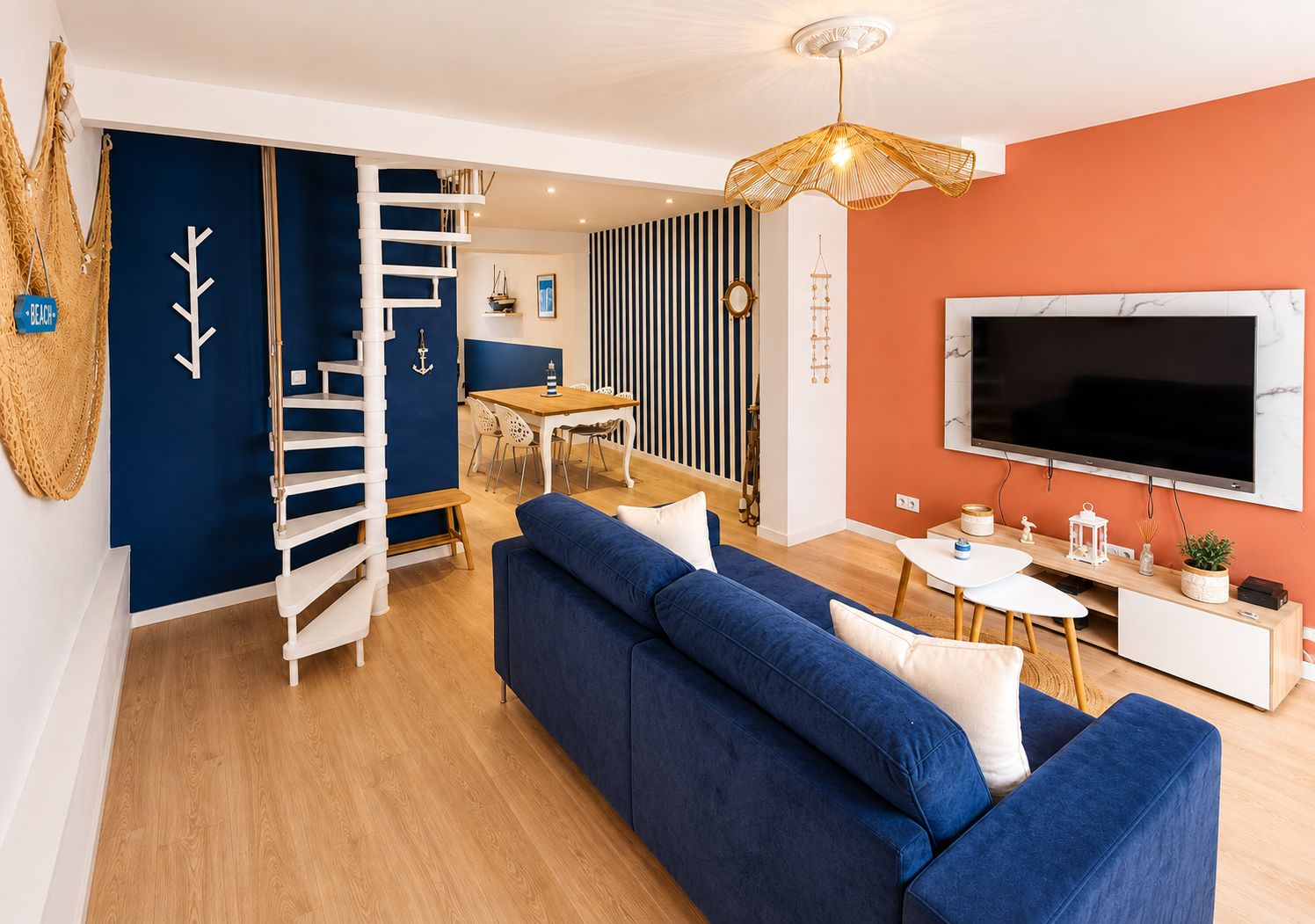 Un salon moderne avec un canapé bleu, un escalier en bois, un coin repas et un téléviseur fixé au mur.