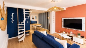 Ein modernes Wohnzimmer mit einem blauen Sofa, einer Holztreppe, einem Essbereich und einem an der Wand befestigten Fernseher.
