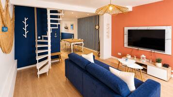 Un salon moderne avec un canapé bleu, un escalier en bois, un coin repas et un téléviseur fixé au mur.