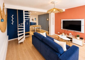 Ein modernes Wohnzimmer mit einem blauen Sofa, einer Holztreppe, einem Essbereich und einem an der Wand befestigten Fernseher.