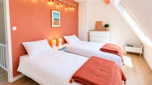 Ein Schlafzimmer mit zwei Betten, einer weißen Kommode, einem kleinen Tisch und einer Pflanze. Die Decke ist schräg und verfügt über ein Oberlicht.