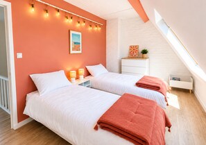 Ein Schlafzimmer mit zwei Betten, einer weißen Kommode, einem kleinen Tisch und einer Pflanze. Die Decke ist schräg und verfügt über ein Oberlicht.