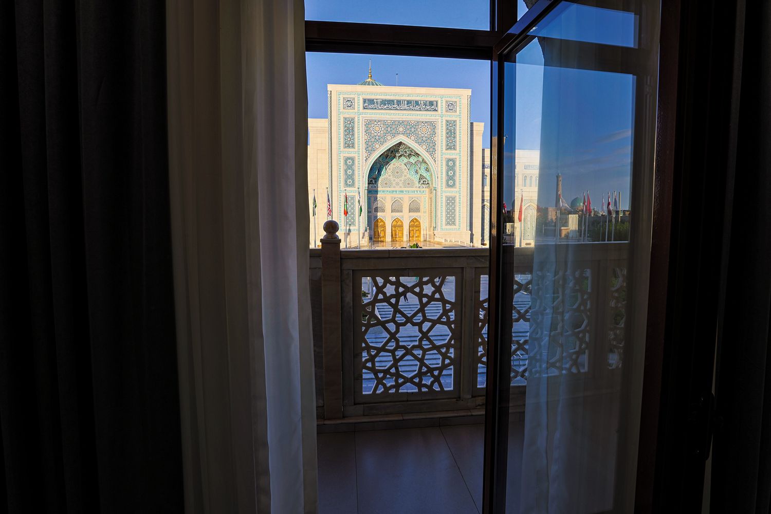 Vista a través d'una finestra amb una reixa decorativa, que mostra un edifici amb un gran arc i treballs de rajola intricats.