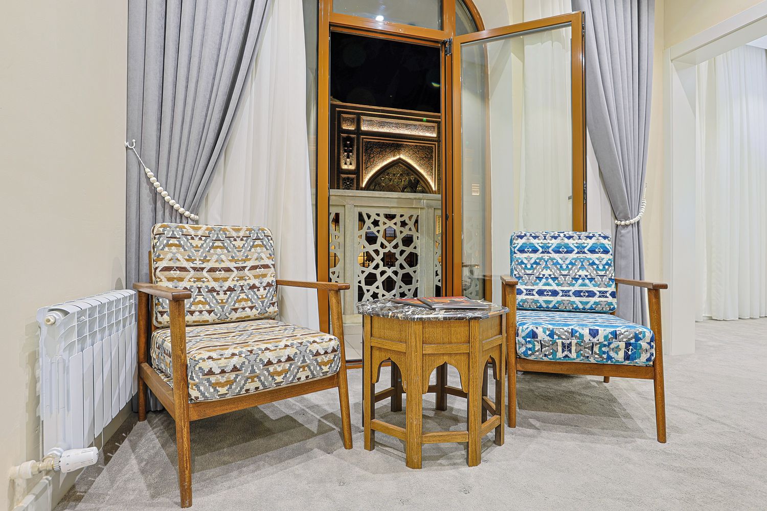 Two patterned chairs, a small wooden table, and a decorative mirror in a room with a radiator and curtains.