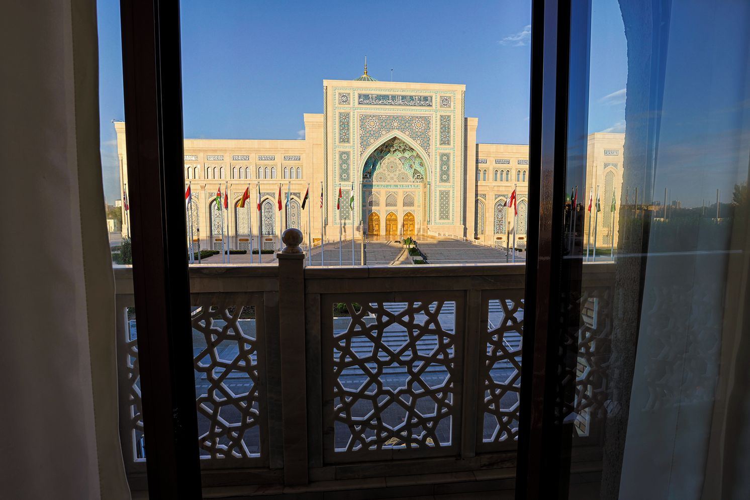 A view of a building with a large archway and multiple flags, viewed through a window with a lattice design.