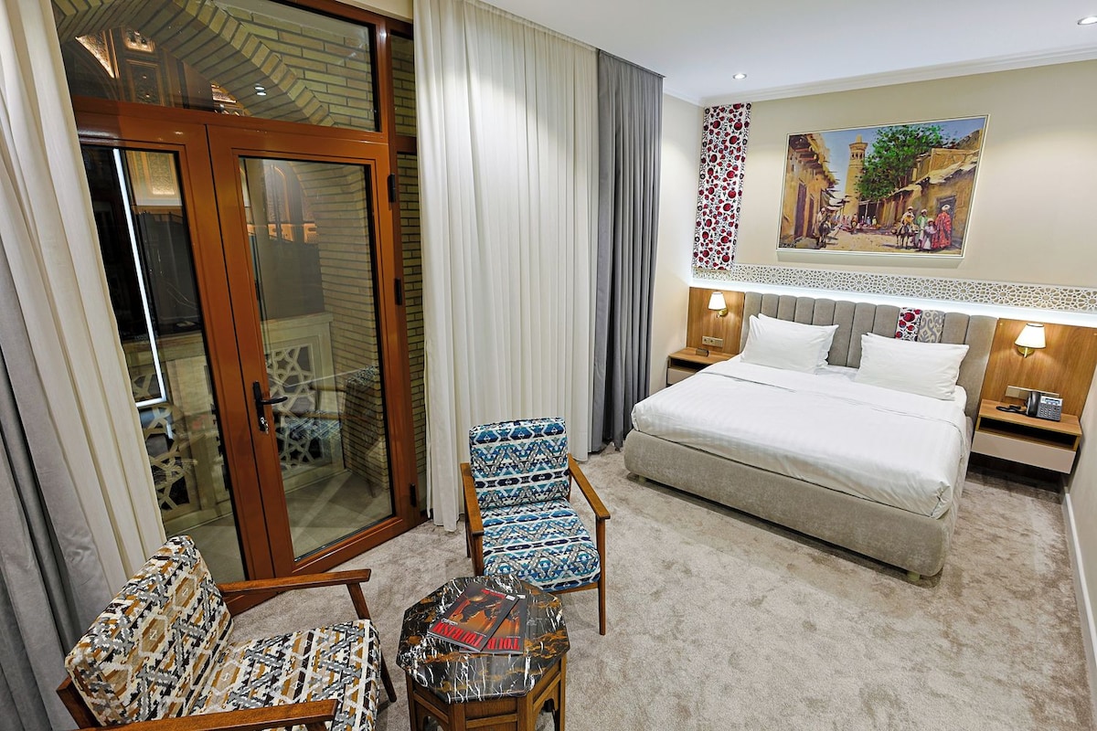 A hotel room with a bed, a chair, a small table, a nightstand, and a painting on the wall.