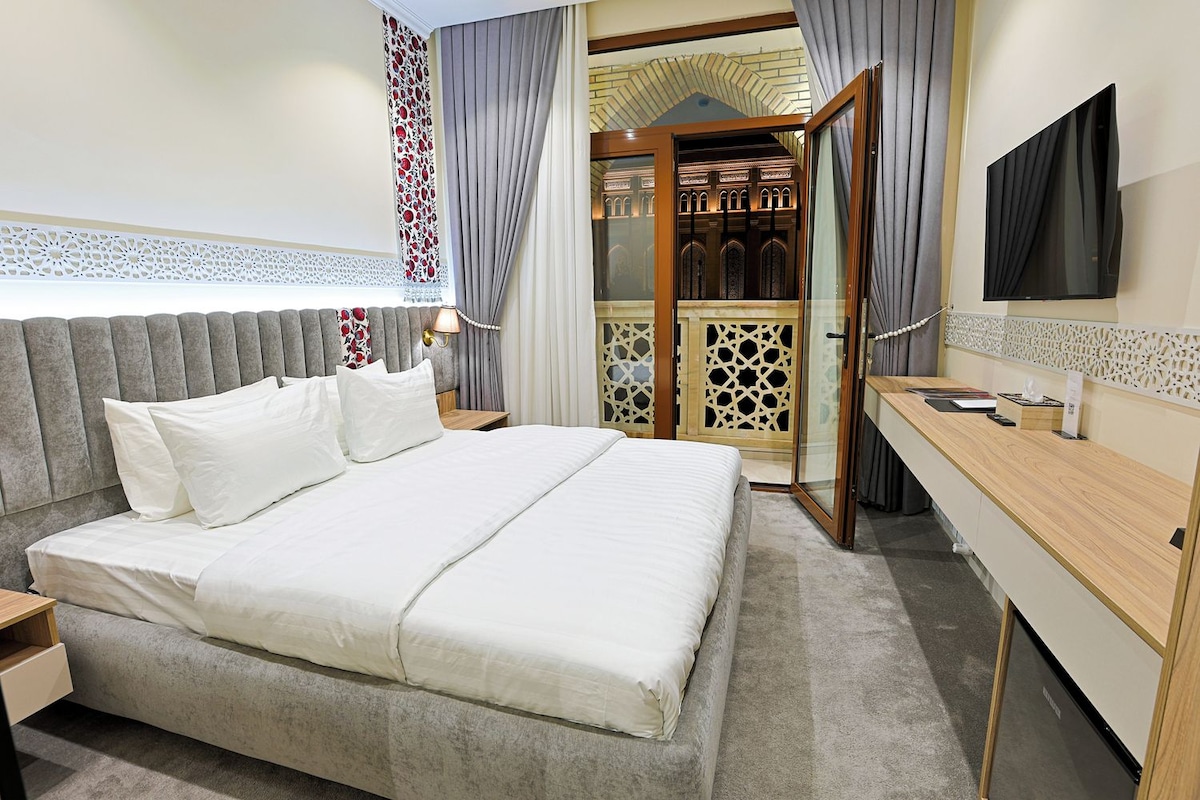 A hotel room with a large bed, a desk, a TV, and a view of a building through a glass door.