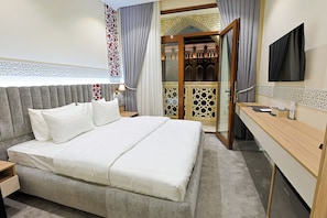 A hotel room with a large bed, a desk, a TV, and a view of a building through a glass door. - Silk Way Hotel Tashkent by HMG (Tashkent)