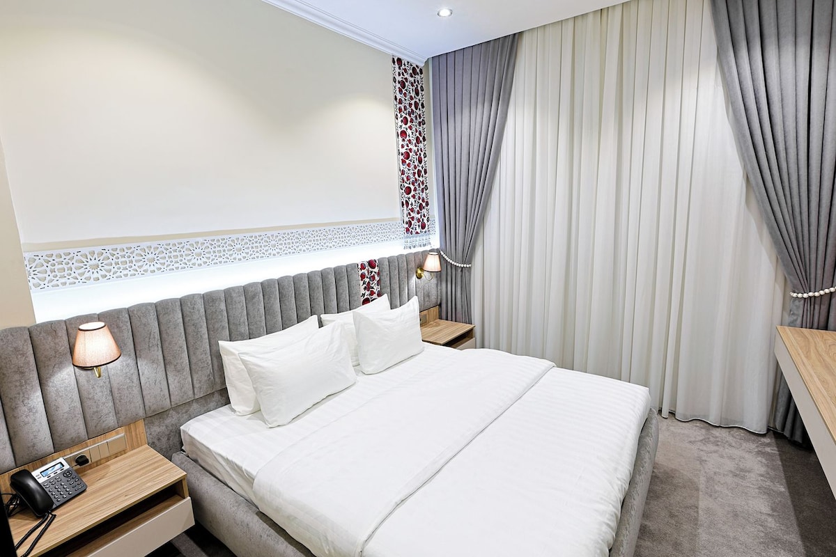 A hotel room with a large bed, bedside lamps, a telephone, a decorative wall panel, and a window with curtains.
