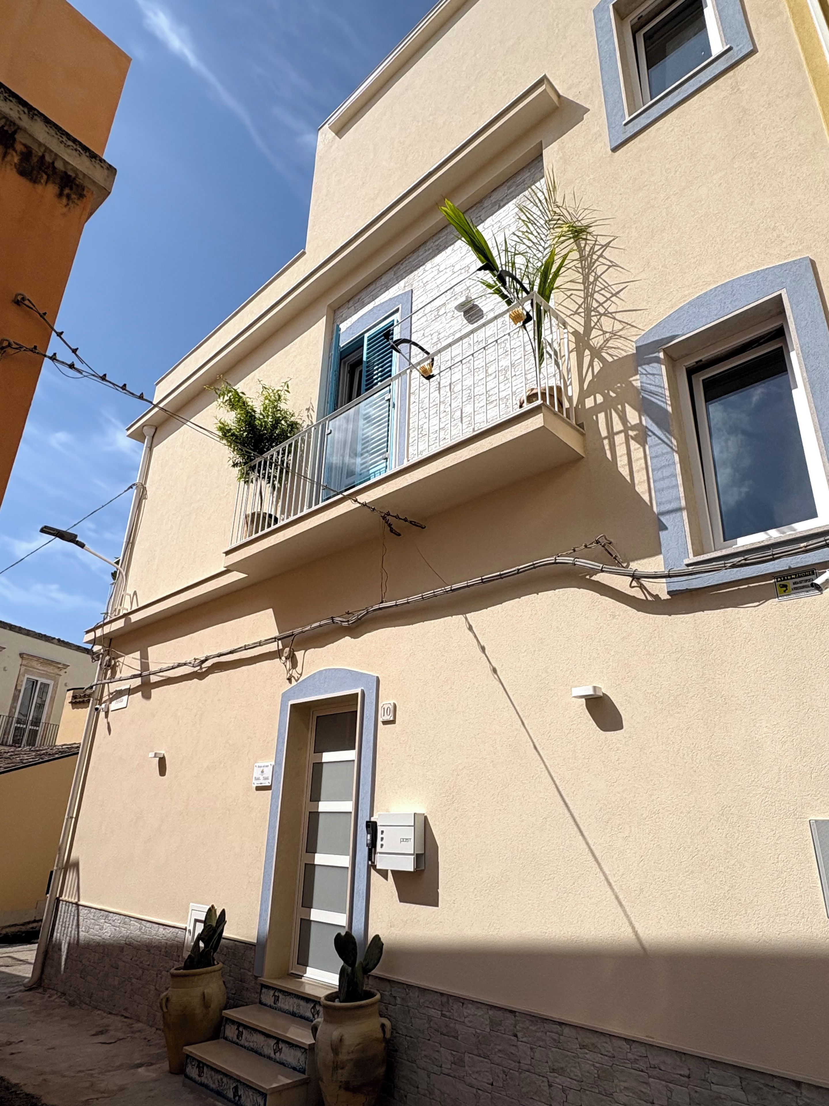 Un bâtiment à deux étages doté d’un balcon, d’une porte et d’un petit jardin avec des plantes.