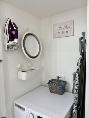 A laundry room with a white washing machine, a hanging iron, a mirror, and a sign that reads 'Welcome to the Laundry Room.'