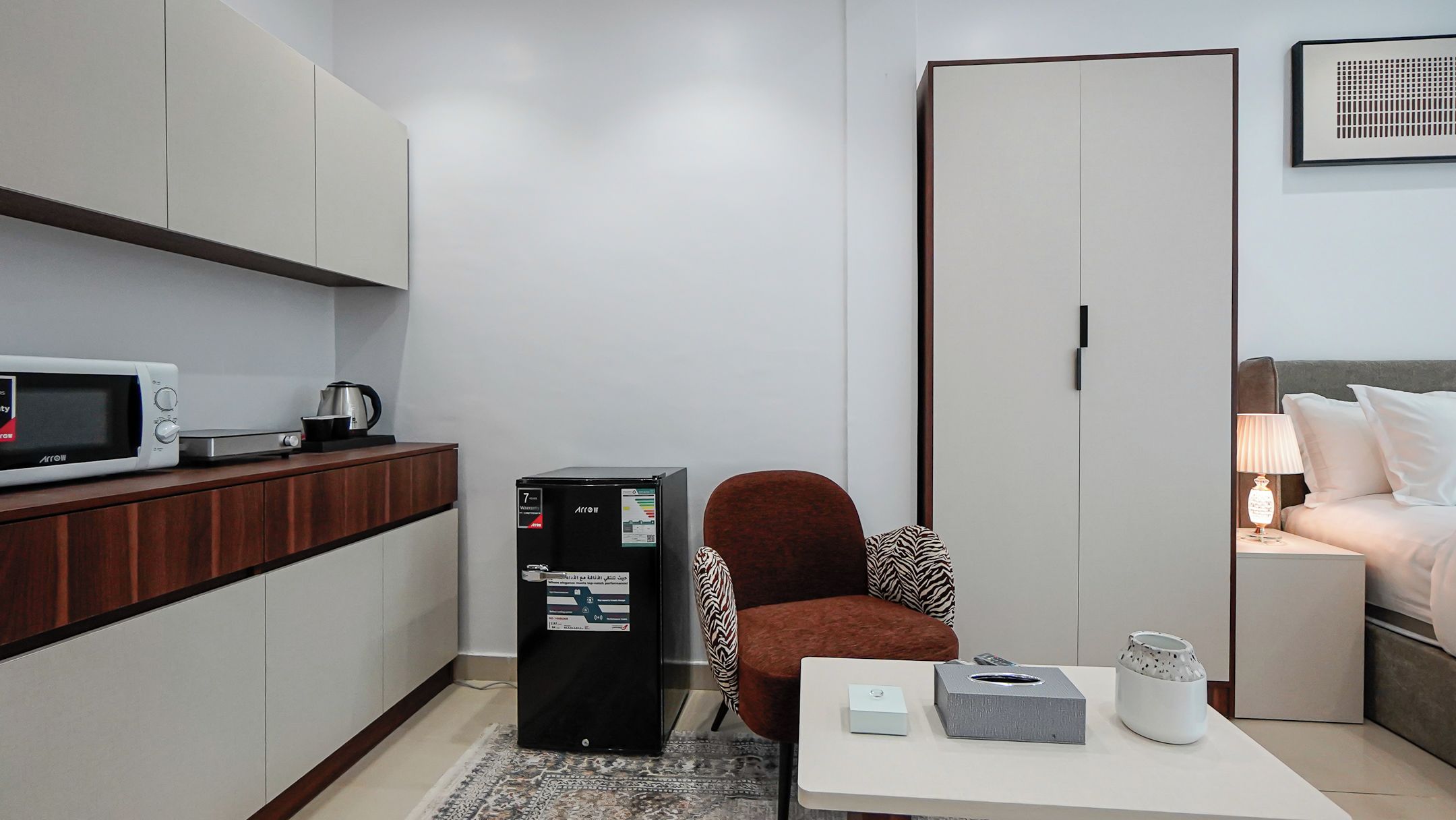 Habitación de hotel compacta con kitchenette, una cama, un armario y una pequeña mesa con un control remoto.