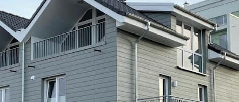 Un edificio residencial moderno de dos pisos con fachada gris, un balcón en el nivel superior y una puerta de garaje visible.