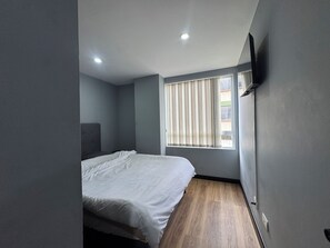 A bedroom with a bed, a window with blinds, a flat-screen TV mounted on the wall, and a ceiling light.