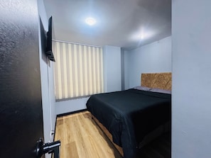 A small, single-bed room with a wooden floor, a black bedspread, a ceiling-mounted TV, and a window with vertical blinds.