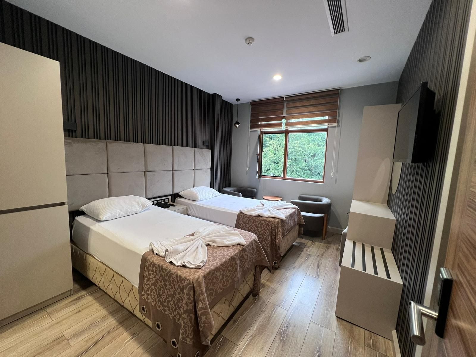 Een hotelkamer met twee bedden, een tafeltje, een stoel, een televisie en een raam met uitzicht op groen.