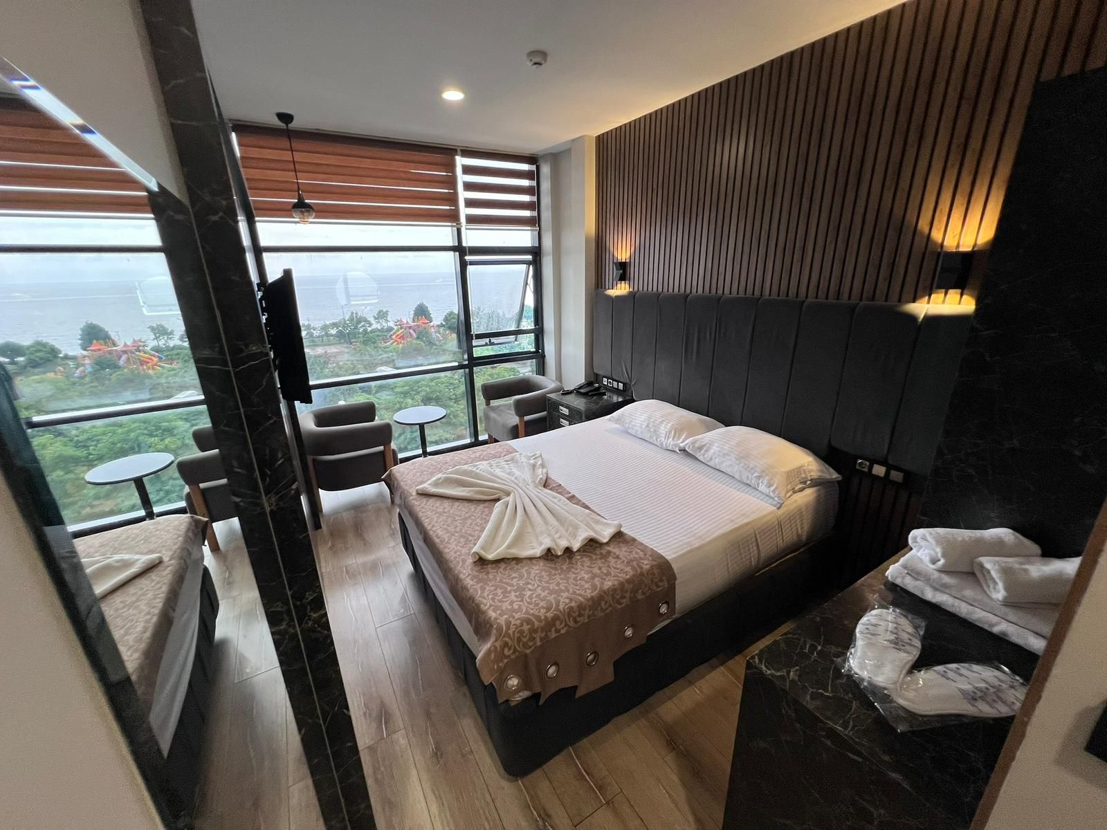 Una habitación de hotel moderna con una cama grande, una zona de estar con dos sillones y una mesita, y vistas al exterior a través de amplios ventanales.