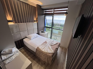 Une chambre moderne avec un grand lit, une table de chevet, une commode, un téléviseur et une fenêtre donnant sur l’extérieur.
