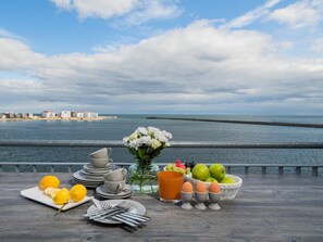 Ein Frühstückstisch mit Tellern, Tassen, Zitronen, Eiern und einem Blumenstrauß, mit Blick auf ein Gewässer und Gebäude.
