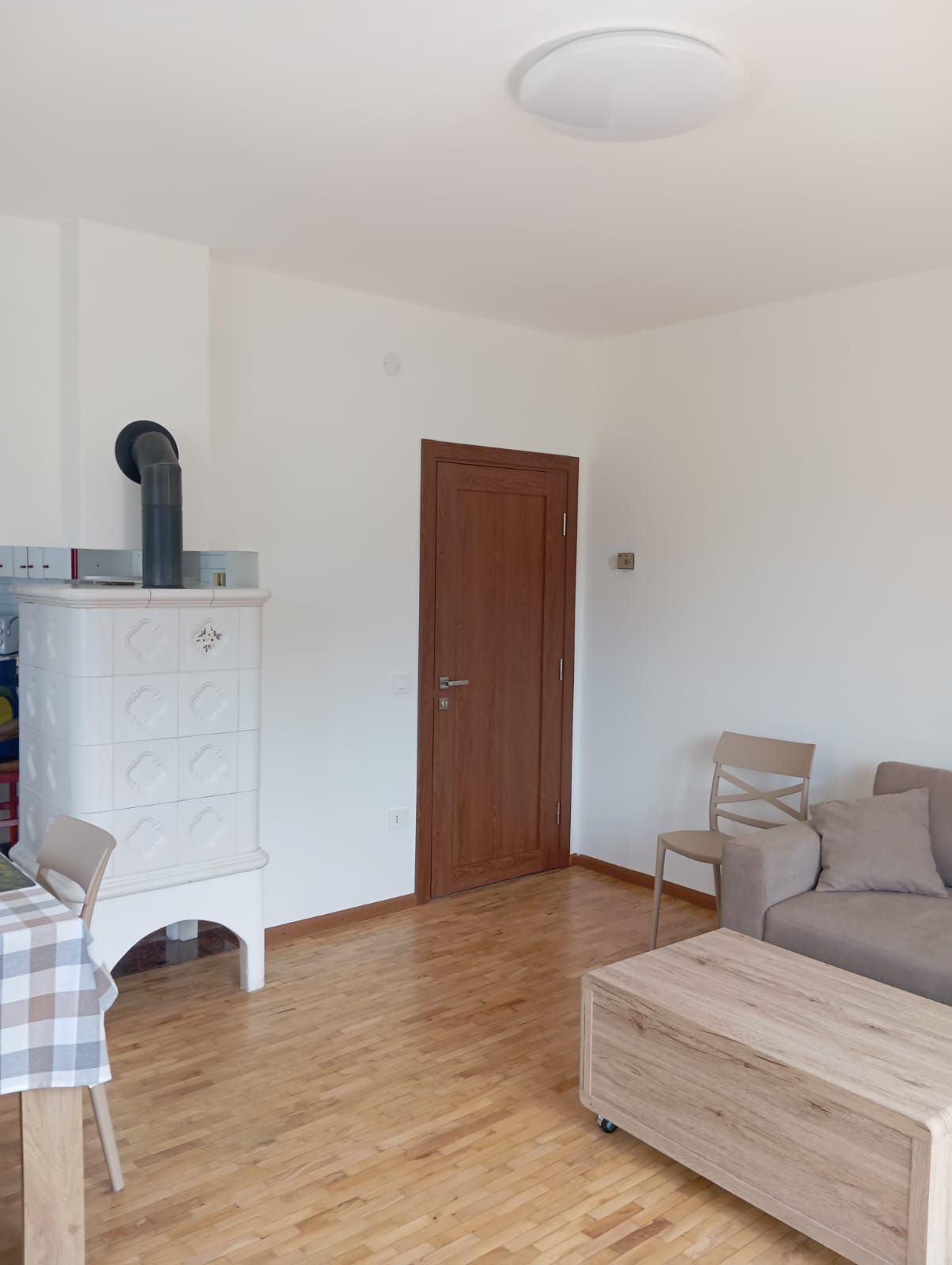 Una stanza con pavimento in legno, una porta marrone, un armadietto bianco con un oggetto nero sopra, un divano beige e un tavolo a scacchi con una sedia.