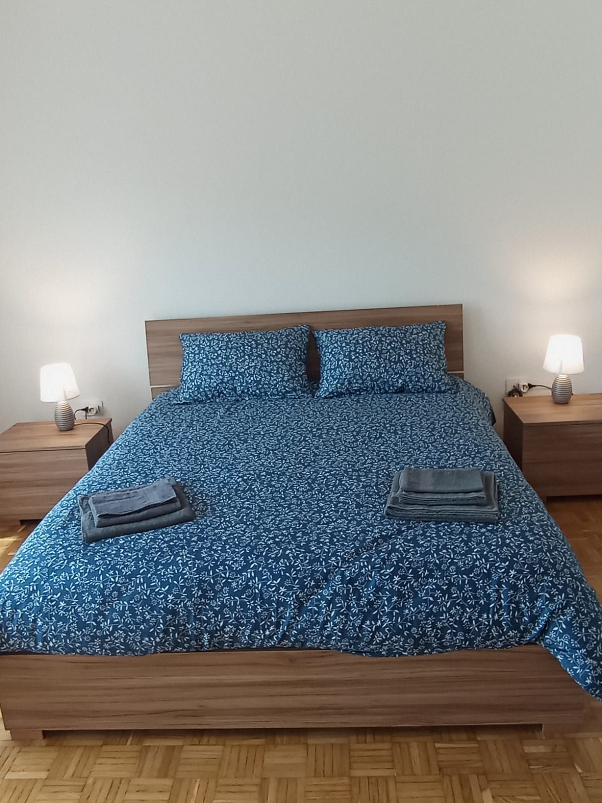 A bed with a floral patterned bedspread, two pillows, two bedside tables with lamps, and a wooden floor.