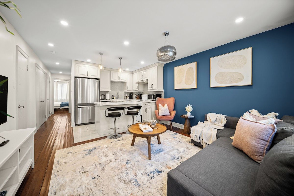 A modern kitchen with stainless steel appliances, a dining area with bar stools, a living room with a sofa and a coffee table, and a blue accent wall.