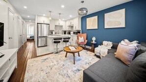 Una cocina moderna con electrodomésticos de acero inoxidable, un comedor con taburetes de bar, una sala de estar con sofá y una mesa de centro, y una pared decorativa azul.