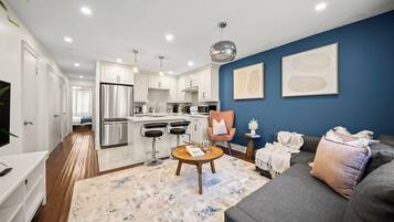 A modern kitchen with stainless steel appliances, a dining area with bar stools, a living room with a sofa and a coffee table, and a blue accent wall.