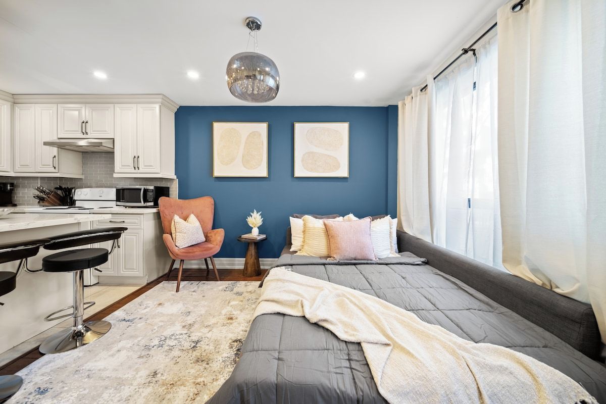 A modern bedroom with a bed, a desk, and a chair. There is a kitchen area with cabinets and appliances.