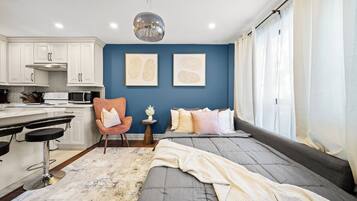 A modern bedroom with a bed, a desk, and a chair. There is a kitchen area with cabinets and appliances.