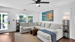 A modern living room with a sectional sofa, a ceiling fan, a large window with blinds, and a painting on the wall. - **SUNSET ON FOGARTY @ MIDTOWN** Newly Renovated Home & Pool + Last Key White Glo (Key West)
