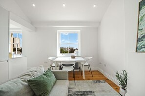 A modern, well-lit room with a white sofa, a dining table with chairs, a large window with a city view, and a small plant. - Between Camden & Kings Cross. Above the Ordinary - Pass the Keys (London)