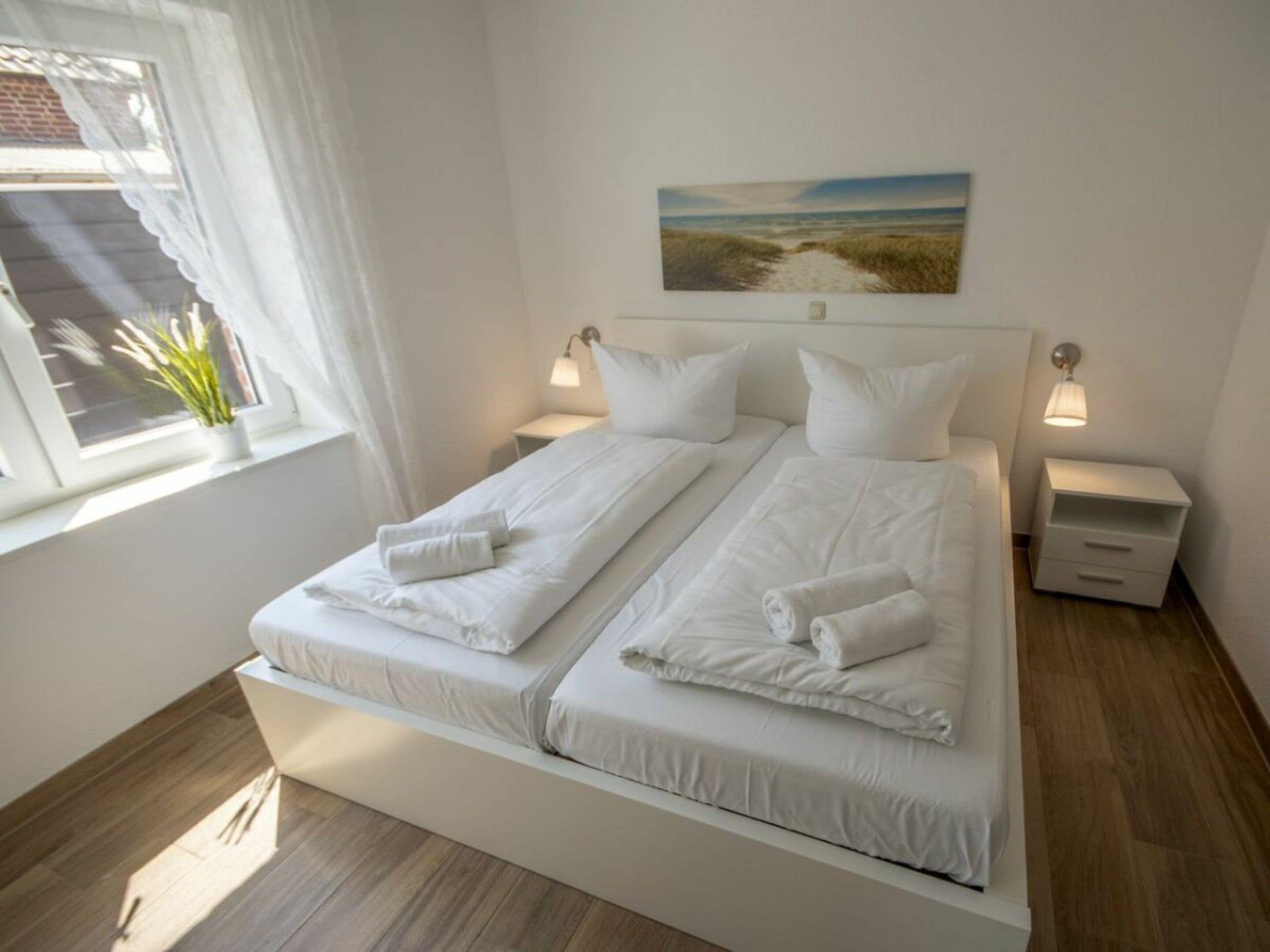 Een tweepersoonsbed met wit beddengoed en kussens, een nachtkastje met een lamp, en een schilderij van een strandgezicht aan de muur.