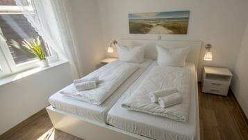 Een tweepersoonsbed met wit beddengoed en kussens, een nachtkastje met een lamp, en een schilderij van een strandgezicht aan de muur.