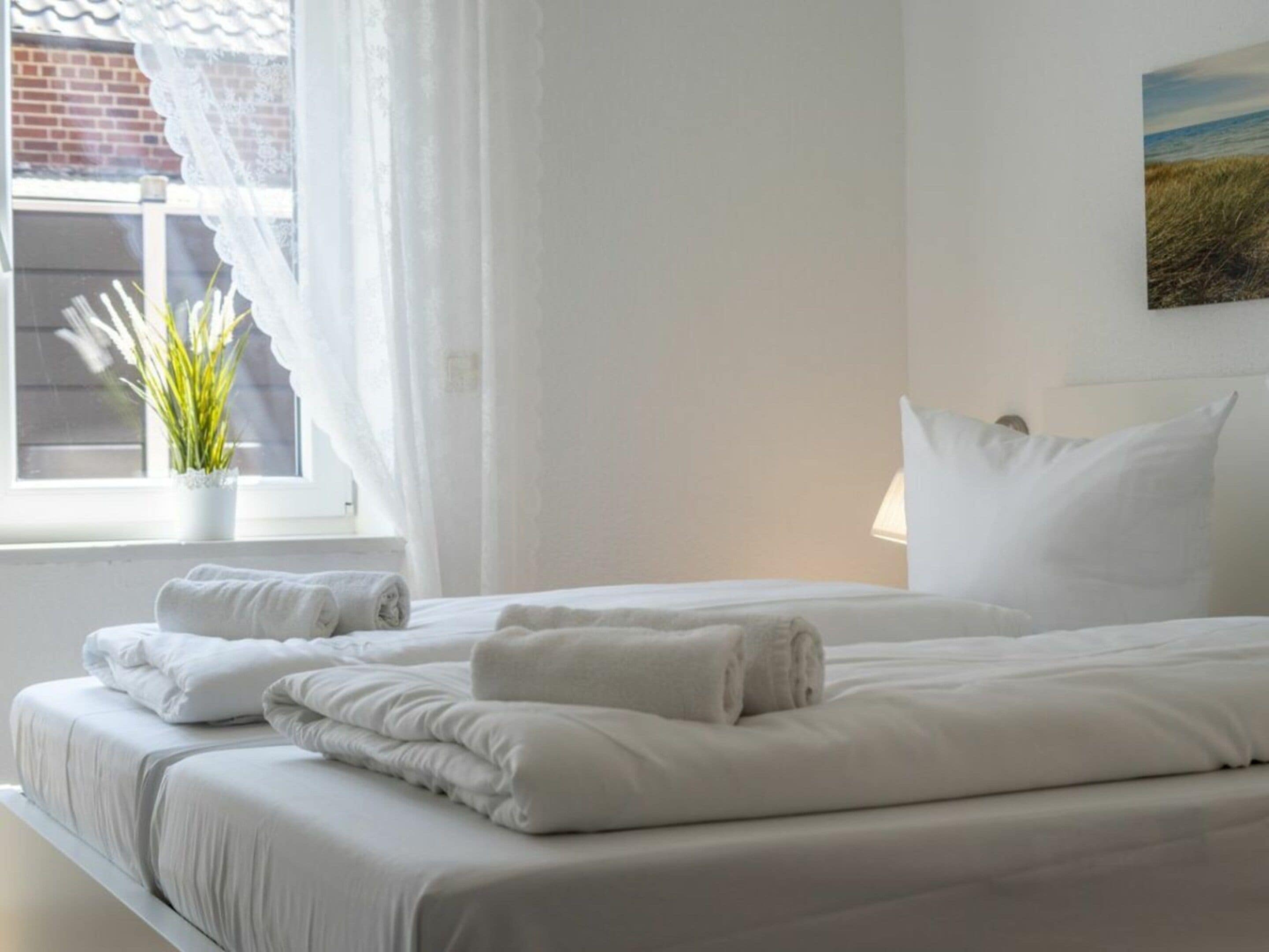 Ein Hotelzimmer mit zwei Betten, weißer Bettwäsche, einem Fenster mit leichtem Vorhang, einer Topfpflanze und einem Bild an der Wand.