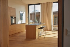 Eine moderne Küche mit Holzausbau, einer zentralen Kochinsel, Barhocker und einem großen Fenster mit Vorhängen.