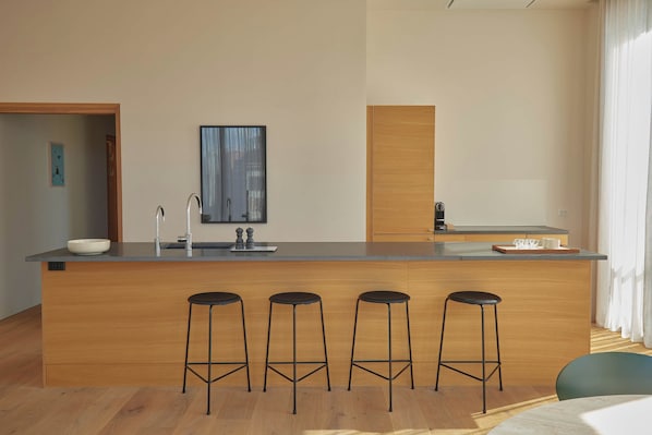 A modern kitchen with a central island, bar stools, a coffee machine, and a large window.
