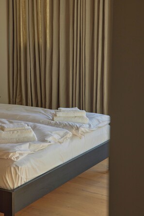 A bed with white linens and folded towels, a wooden floor, and beige curtains.