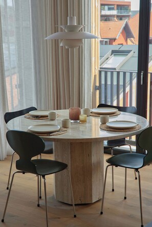 Uma sala de jantar com uma mesa redonda, quatro cadeiras, um lustre e vista para prédios pela janela.