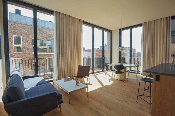 Un salon moderne avec un canapé, une table basse et un coin repas. De grandes fenêtres offrent une vue sur le paysage urbain.