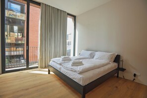 Une chambre à coucher avec un grand lit, une fenêtre avec des rideaux, un plancher en bois et une vue sur un bâtiment à l’extérieur.