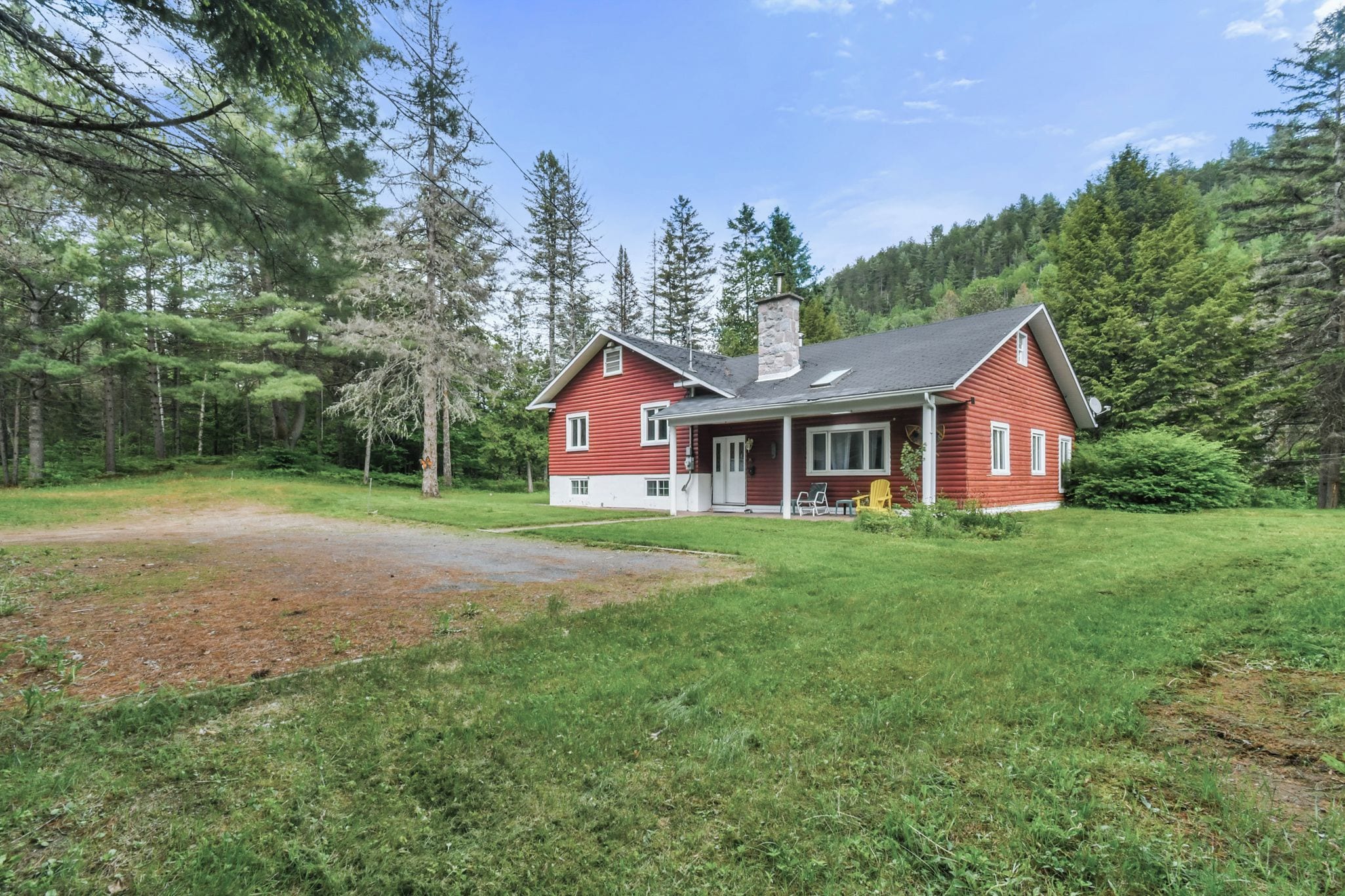A red log cabin with a chimney, surrounded by a grassy yard and dense forest.