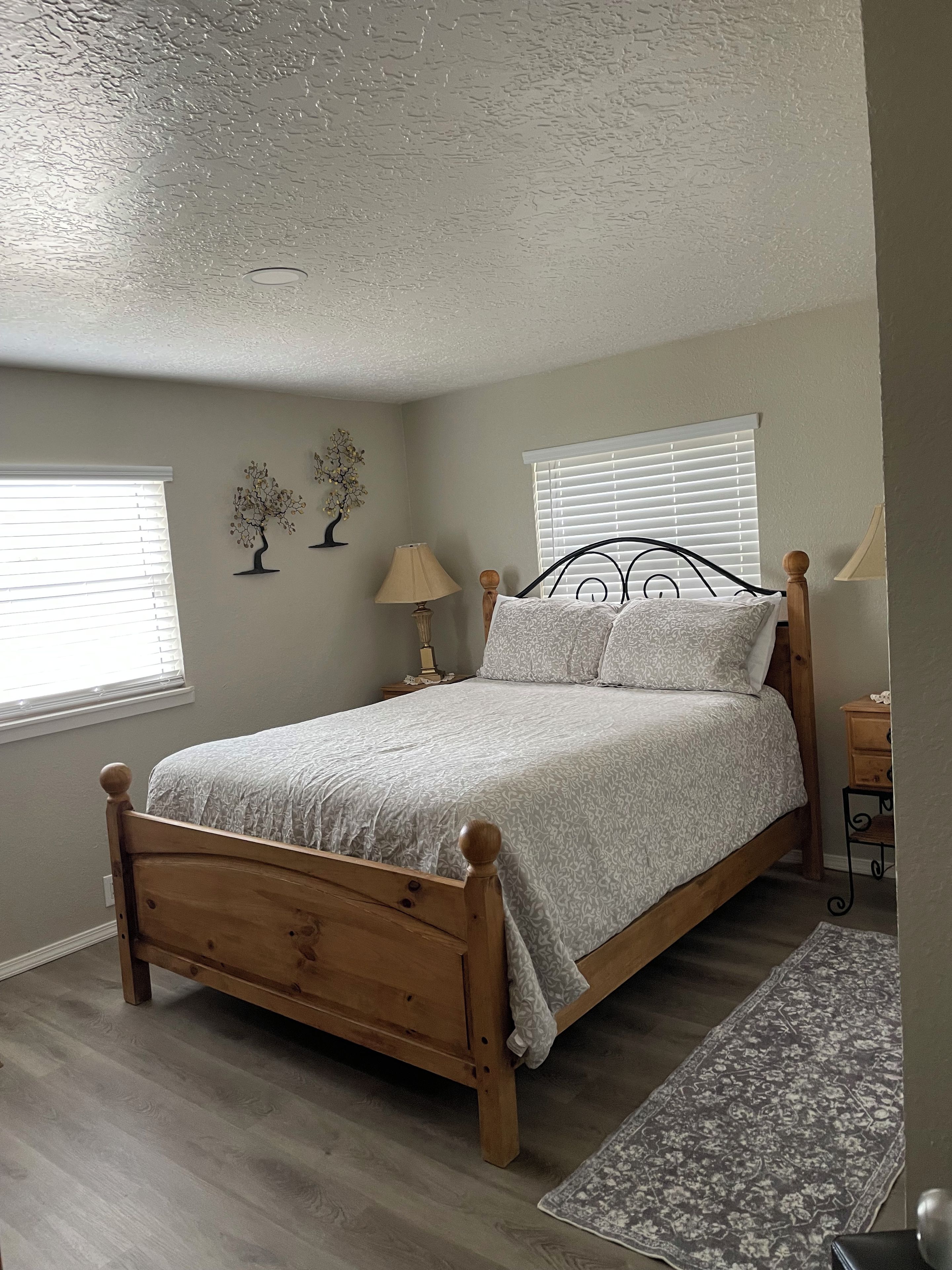 A bedroom with a wooden bed, two bedside lamps, a window with blinds, and a decorative tree on the wall.