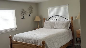 A bedroom with a wooden bed, two bedside lamps, a window with blinds, and a decorative tree on the wall.