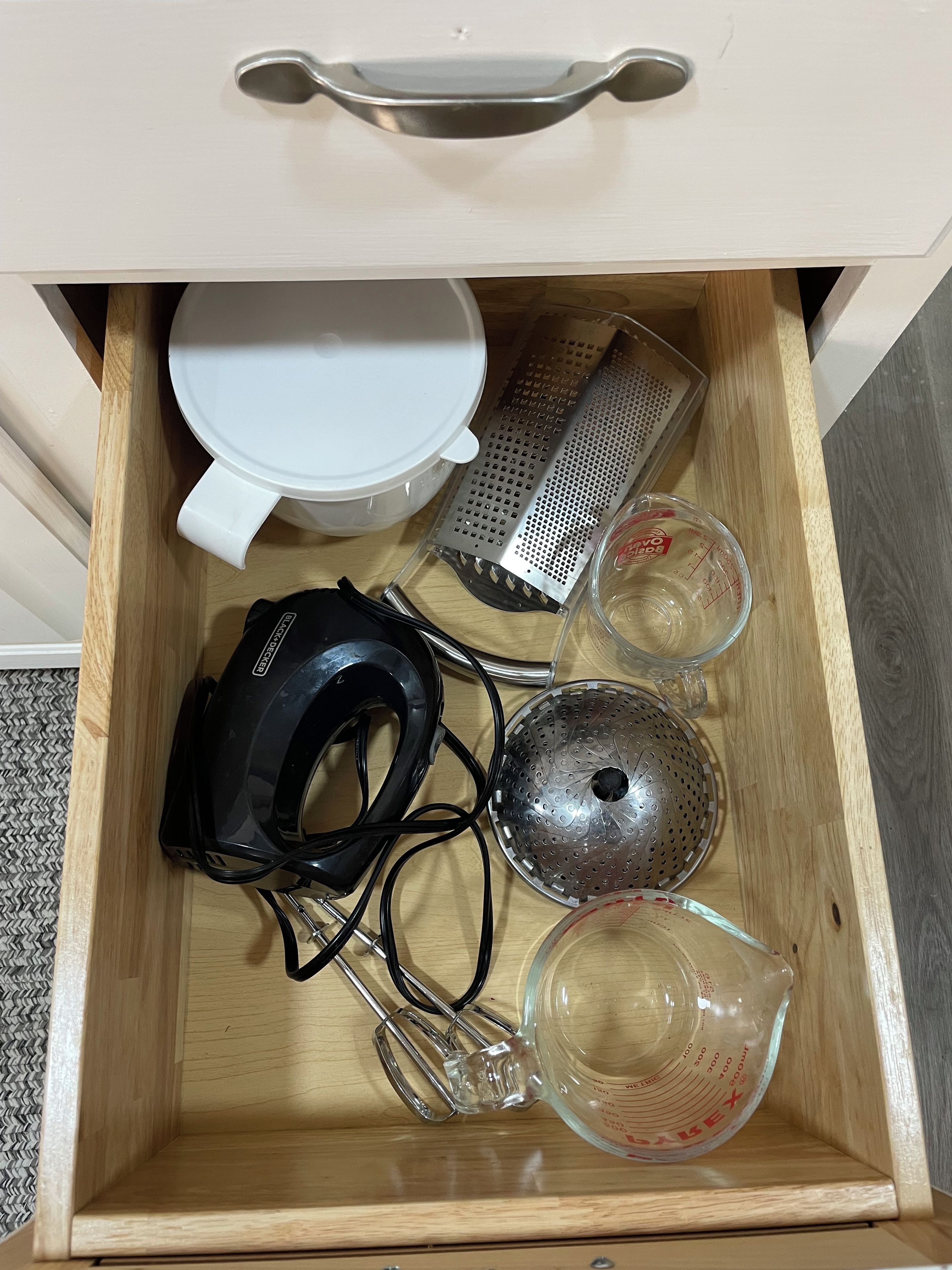 A drawer containing kitchen utensils including a grater, measuring cups, and a whisk.
