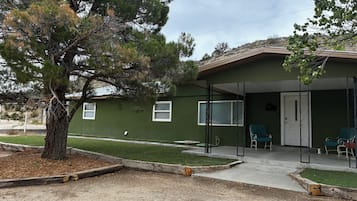 Um edifício com fachada verde, varanda coberta com cadeiras e uma árvore em primeiro plano.