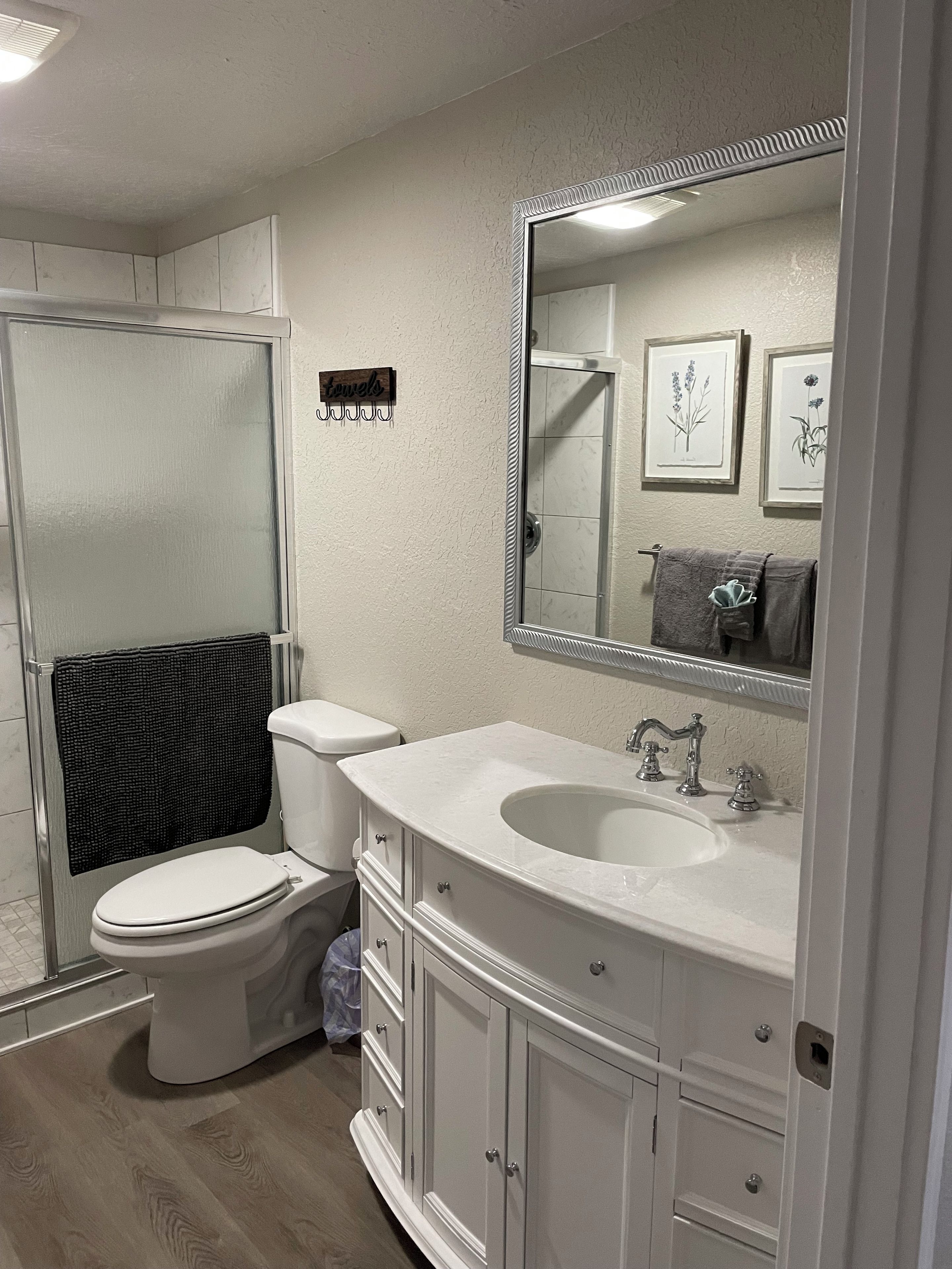 A bathroom with a toilet, sink, and shower. There is a large mirror above the sink and framed botanical prints on the wall.