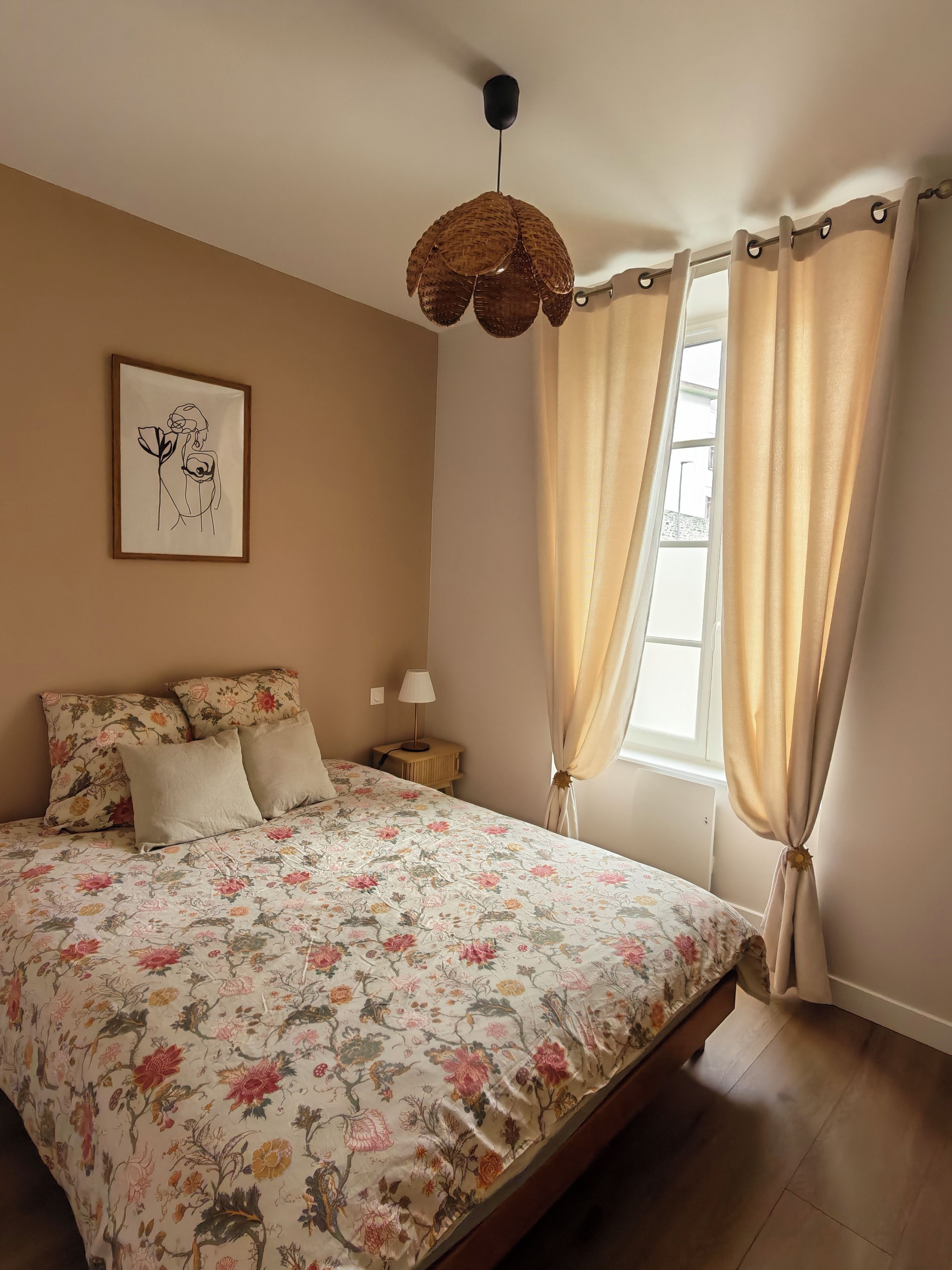 A bedroom with a floral bedspread, a hanging light fixture, a bedside table with a lamp, and a window with curtains.