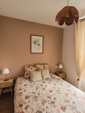 A bedroom with a floral bedspread, two bedside lamps, a hanging woven light fixture, and a framed artwork on the wall. - Suite Adélaïde, Élégance Rétro au cœur de la ville (Landerneau)